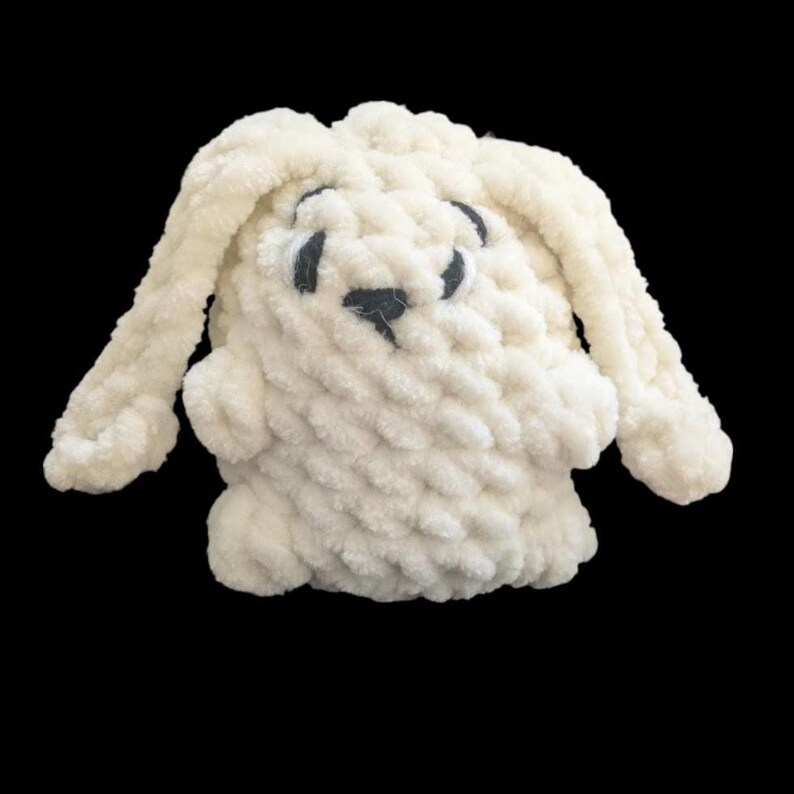 May include: A cream-coloured plush toy shaped like a rabbit, crafted from thick, chunky yarn with a soft, textured appearance. The stuffed animal features long, floppy ears, two dark, oval-shaped eyes, and a small, dark, triangular nose.