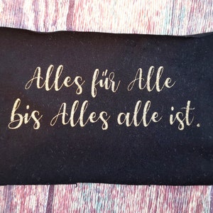 Könnte beinhalten: Schwarzer Stoff-Patch mit goldener Aufschrift "Alles für Alle bis Alles alle ist."