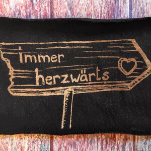 Könnte beinhalten: Ein schwarzer Stoff-Patch mit einem goldfarbenen Holzschild, auf dem "Immer Herzwärts" mit einem Herzsymbol steht. Das Schild zeigt nach rechts.