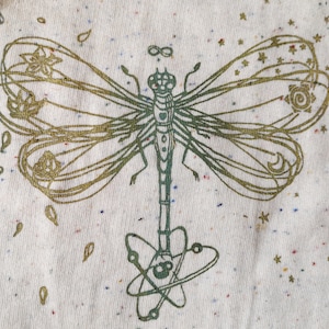 Könnte beinhalten: Ein weißer Stoff mit einem goldenen und grünen Libellen-Design. Die Libelle hat ein Unendlichkeitszeichen über ihrem Kopf und ein Atomzeichen unter ihrem Körper. Der Stoff ist mit kleinen, bunten Punkten bedeckt.