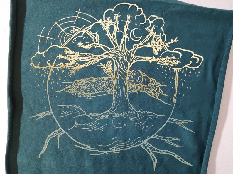 Pu&ograve; includere: Un disegno dorato dell'albero della vita su uno sfondo di tessuto blu scuro. L'albero ha rami che raggiungono una nuvola con gocce di pioggia che cadono da essa. Le radici dell'albero si estendono verso il basso fino a una linea ondulata che rappresenta l'acqua.