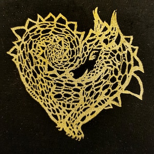 Könnte beinhalten: Ein goldfarbenes herzförmiges Design mit einem wirbelnden Muster, das an einen Drachen oder eine mythische Kreatur erinnert, auf schwarzem Hintergrund.