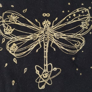 Könnte beinhalten: Schwarzer Stoff mit einem goldfarbenen Libellen-Design. Die Libelle hat detaillierte Flügel mit floralen und himmlischen Elementen. Kleine Sterne und tropfenförmige Formen umgeben die Libelle. Das Design hat eine aufwendige, handgezeichnete Ästhetik.
