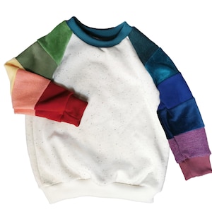 Könnte beinhalten: Ein weißes Sweatshirt mit gesprenkelten Details und einem türkisfarbenen Ausschnitt. Die Ärmel sind ein Patchwork aus Regenbogenfarben, darunter Grün, Gelb, Rot, Blau und Lila. Die Bündchen sind einfarbig rot und lila.