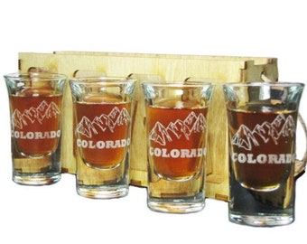 May include: A set of four shot glasses filled with a brown liquid, each etched with a mountain range design and the word "COLORADO". The glasses are held in a light-colored wooden carrier.