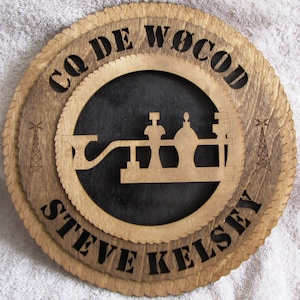 May include: A wooden sign with a brown stain and a black center. The sign has a rope border and the text "CQ DE WOCOD STEVE KELSEY" in black lettering. The center of the sign has a silhouette of a radio transmitter.