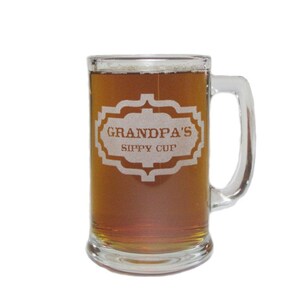 May include: A clear glass beer mug with a handle. The mug is filled with a dark brown liquid. The mug is etched with the words "Grandpa's Sippy Cup".