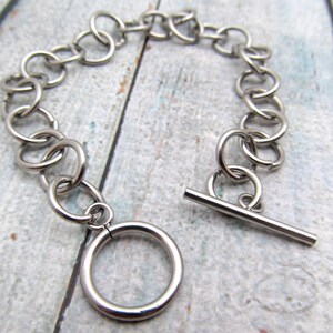 May include: A silver chain bracelet with a t-bar clasp. The chain is made of interlocking circles.