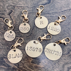 May include: Seven gold-coloured metal keychains with different engraved text. The keychains feature a circular tag with a split ring and a lobster claw clasp. The text on the tags reads: "K&L", "250", "R", "1220", "13579", "SLAM", and "TRUTH".
