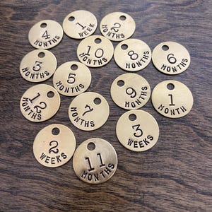 Peut inclure: Un ensemble de 13 jetons en laiton avec des chiffres et les mots "mois" ou "semaines" gravés dessus. Les jetons sont disposés sur une surface en bois.