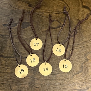 Pode incluir: Um conjunto de seis etiquetas circulares de metal dourado, cada uma suspensa por um fino cordão de couro castanho. As etiquetas estão estampadas individualmente com os números pretos 12, 14, 15, 16, 17 e 18. Cada etiqueta tem um pequeno anel.