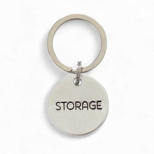 May include: A silver-colored keychain with a round pendant. The pendant has the word "STORAGE" in a bold, sans-serif font. The keychain ring is attached to the pendant with a small loop. The background is plain white.