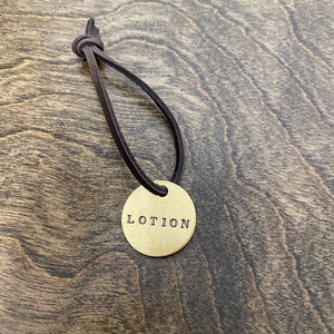May include: A gold-colored metal tag with the word "LOTION" engraved on it. The tag is attached to a brown leather cord.