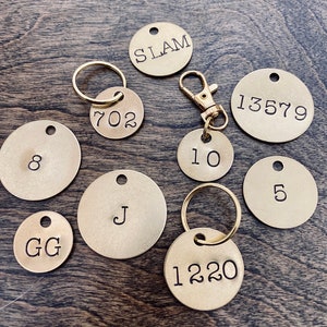 May include: A collection of nine brass keychains with various numbers, letters, and words engraved on them. The keychains are arranged on a wooden surface.