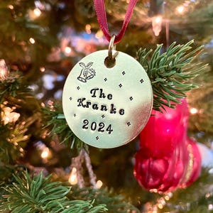 May include: A gold-colored round Christmas ornament with the text "The Kranks 2024" and a small image of a bell with holly leaves.