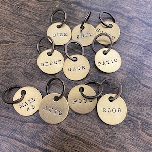 Peut inclure: Un ensemble de dix étiquettes de clés en laiton avec des anneaux noirs. Les étiquettes sont rondes et ont divers mots et chiffres gravés dessus, notamment "Bike", "Shed", "House", "Depot", "Gate", "Patio", "Mail #5", "Auto", "Pool", et "2509".