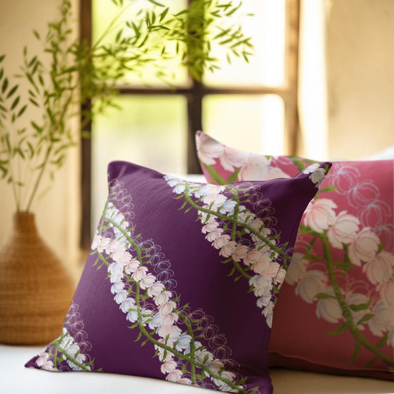 May include: Two decorative throw pillows. One is a deep purple with a floral pattern of white and light pink flowers with green stems. The other pillow is pink with a similar floral design. The pillows are on a white surface.