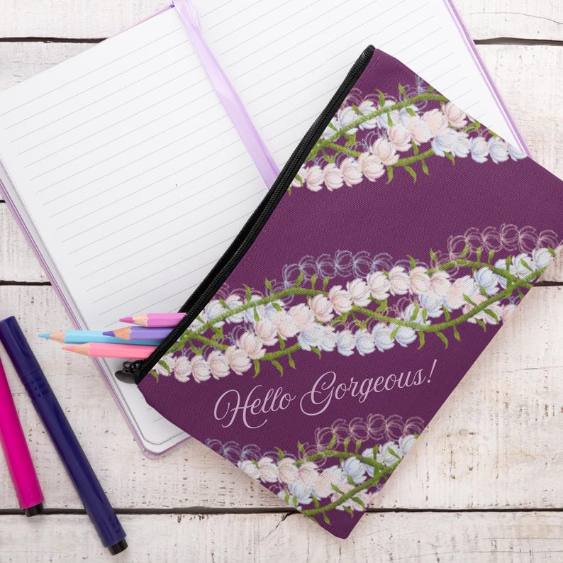 May include: A purple pencil case with a floral design and the text "Hello Gorgeous!". The case is open, revealing colored pencils. A notebook and pens are also visible.