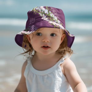 May include: A purple sun hat with a floral design and the name "Lilly" printed on the front. The hat features a wide brim and is worn by a young child on a beach.