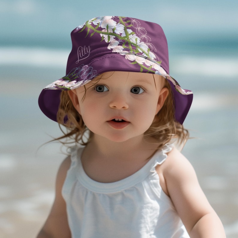 May include: A purple sun hat with a floral design and the name "Lilly" printed on the front. The hat features a wide brim and is worn by a young child on a beach.