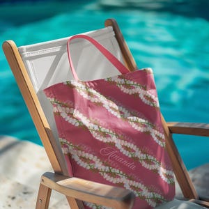 May include: A pink tote bag with a floral pattern of white flowers and green stems, resting on a wooden beach chair. The bag has pink handles and the name "Amanda" is printed on the front.