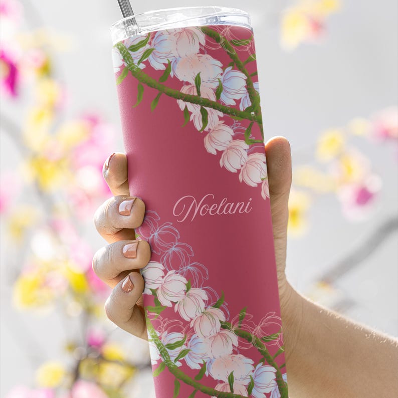 May include: A maroon tumbler with a floral design and the name "Noelani" in cursive. The tumbler features a pattern of white and light blue flowers with green stems. A silver straw is visible at the top.