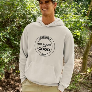 May include: A light grey hooded sweatshirt featuring a black circular graphic on the chest with the text JEREMIAH HIS PLANS ARE GOOD 29:11. The garment includes a front kangaroo pocket, long sleeves, and a drawstring hood, paired with khaki trousers.
