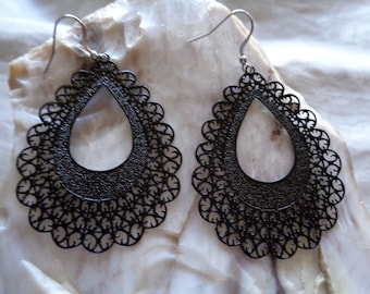 May include: A pair of black filigree teardrop earrings. The earrings have a delicate, openwork design with a scalloped edge. The ear wires are silver-colored. The earrings are displayed on a light-colored, textured surface.