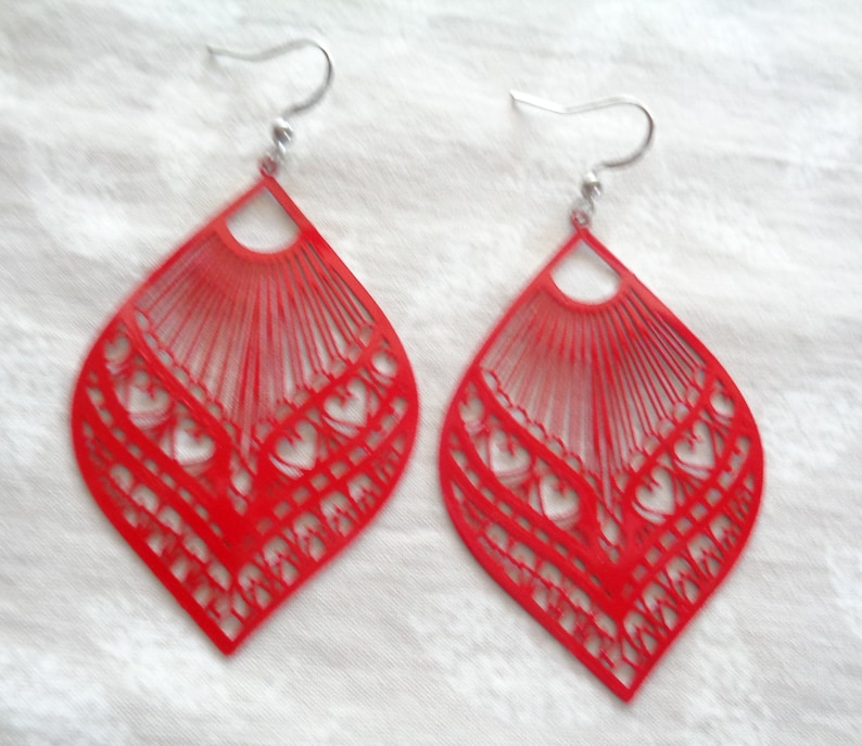 May include: A pair of red, teardrop-shaped earrings. The earrings have a filigree design with heart-shaped cutouts and a fan-like pattern at the top. The ear wires are silver-colored.
