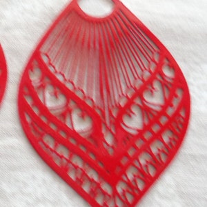 May include: Red, open-work teardrop-shaped earrings. The earrings feature a fan-like design at the top and a heart-shaped pattern at the bottom. The earrings are made of a lightweight material and have a silver-colored hook.