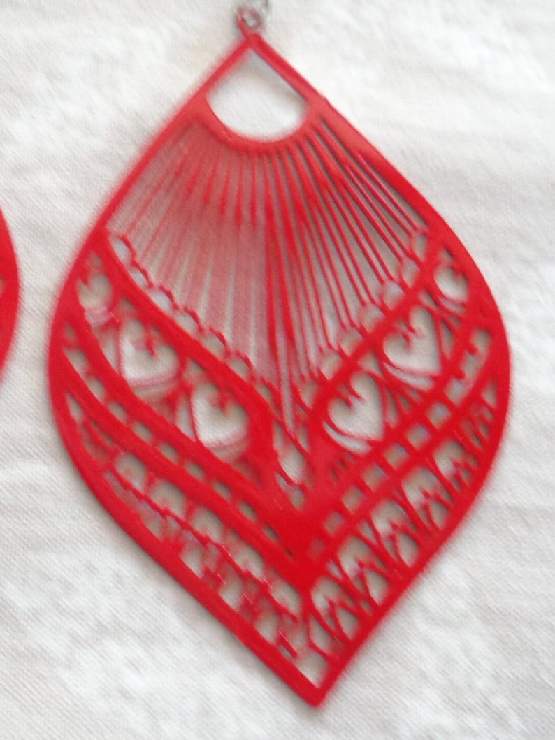 May include: Red, open-work teardrop-shaped earrings. The earrings feature a fan-like design at the top and a heart-shaped pattern at the bottom. The earrings are made of a lightweight material and have a silver-colored hook.