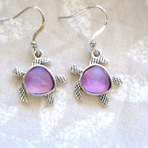 May include: A pair of silver-toned turtle earrings. Each earring features a turtle design with a lavender-colored center. The turtle's body and limbs are detailed with textured patterns, and the earrings have a hook closure.