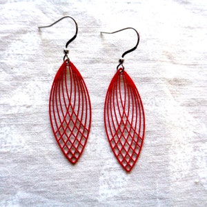 May include: A pair of red, open-work earrings. Each earring is an elongated oval shape with a lattice design. The earrings have black ear wires and silver-colored beads. The earrings are displayed on a white, textured surface.