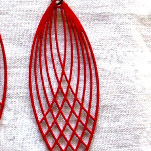 May include: Red, open-work earrings with a geometric design. The earrings are a vibrant red color and feature a series of curved lines that create an elongated, leaf-like shape. The lower portion has a diamond pattern.