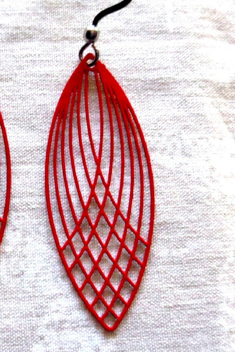 May include: Red, open-work earrings with a geometric design. The earrings are a vibrant red color and feature a series of curved lines that create an elongated, leaf-like shape. The lower portion has a diamond pattern.