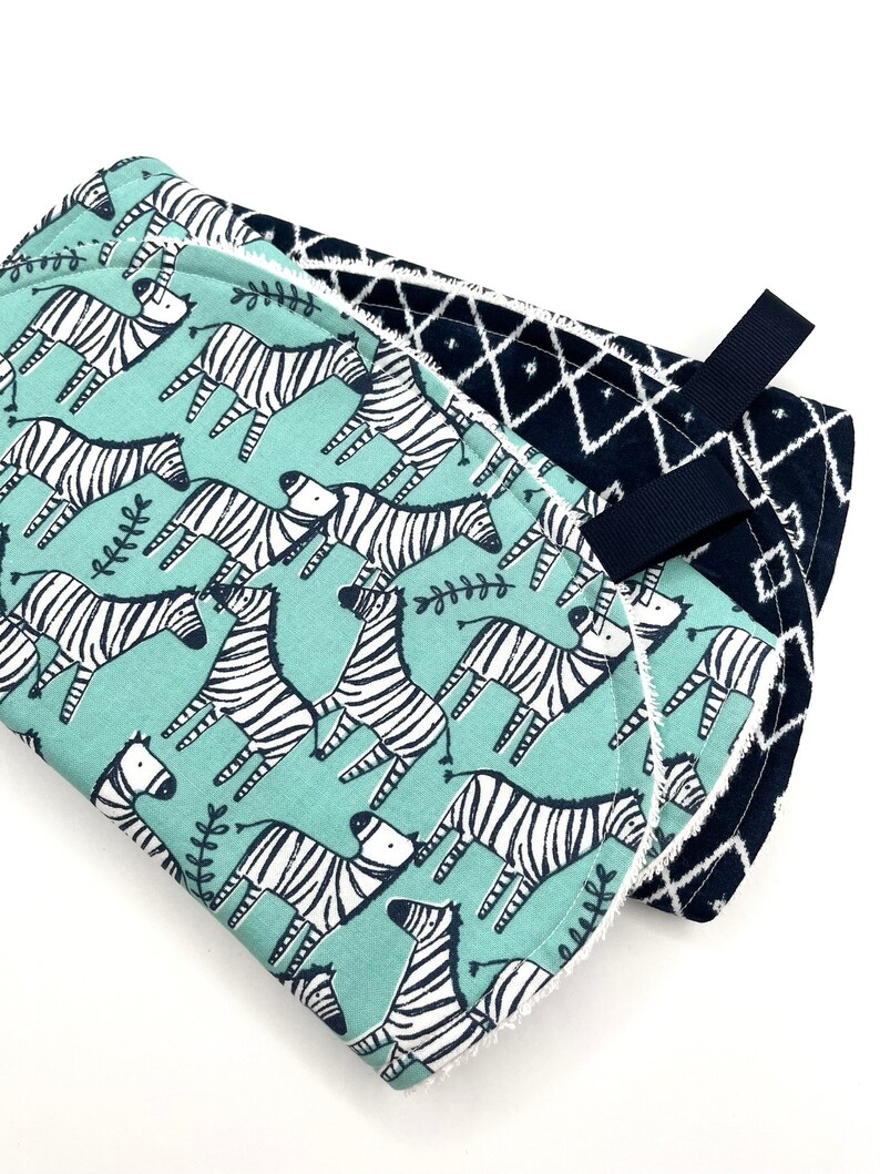 May include: A baby burp cloth with a blue and white zebra print on one side and a black and white geometric pattern on the other side. The cloth has a black strap for easy carrying.