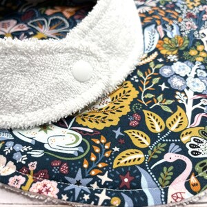 May include: A white terry cloth bib with a snap closure. The bib is lined with a dark blue fabric with a colorful floral pattern featuring birds, stars, and leaves.