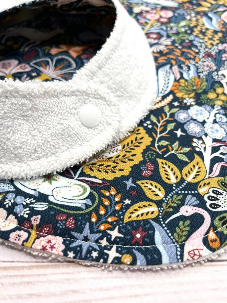May include: A white terry cloth bib with a snap closure. The bib is lined with a dark blue fabric with a colorful floral pattern featuring birds, stars, and leaves.