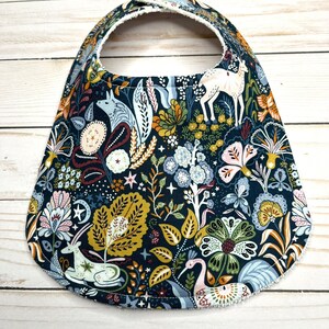 May include: A dark blue baby bib with a whimsical woodland pattern featuring deer, birds, and flowers. The bib has a white fleece lining.