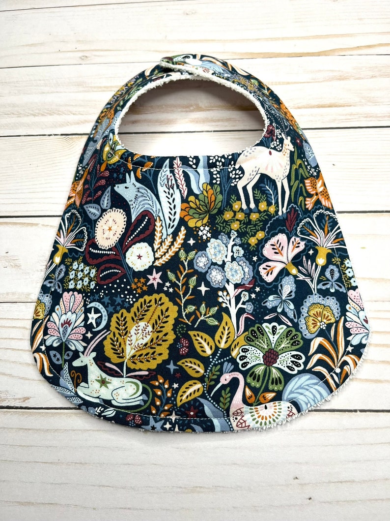 May include: A dark blue baby bib with a whimsical woodland pattern featuring deer, birds, and flowers. The bib has a white fleece lining.