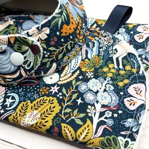 May include: A dark blue fabric bib with a white trim and a floral pattern featuring stars, leaves, and flowers in shades of yellow, green, pink, and blue. The bib has a white button and a navy blue strap.
