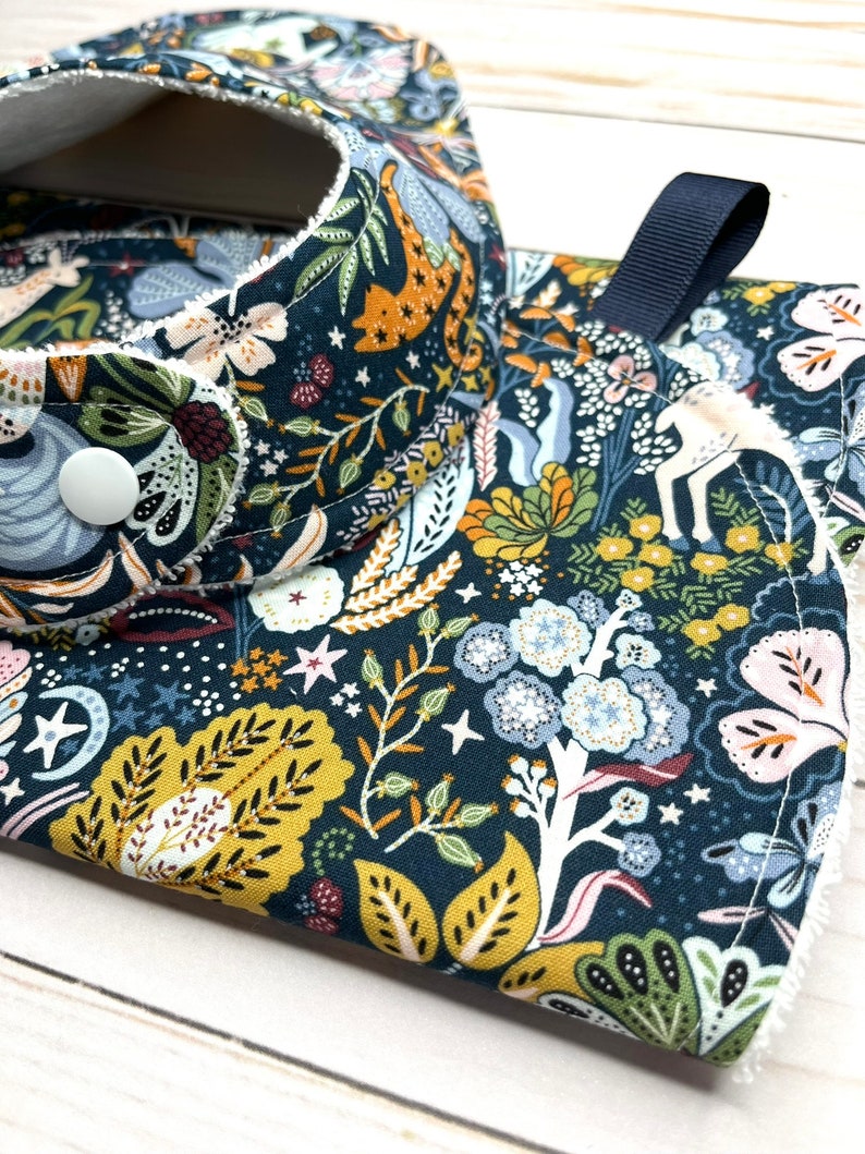 May include: A dark blue fabric bib with a white trim and a floral pattern featuring stars, leaves, and flowers in shades of yellow, green, pink, and blue. The bib has a white button and a navy blue strap.