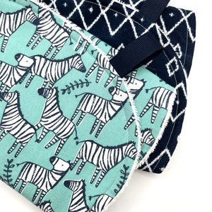 May include: Two baby burp cloths, one with a blue and white geometric pattern and the other with a teal background and black and white zebra print. Both cloths have a white trim and a black ribbon loop.