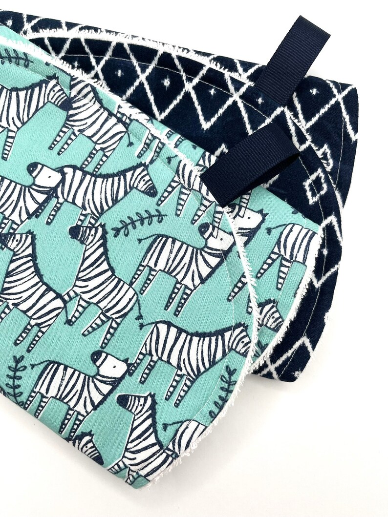 May include: Two baby burp cloths, one with a blue and white geometric pattern and the other with a teal background and black and white zebra print. Both cloths have a white trim and a black ribbon loop.
