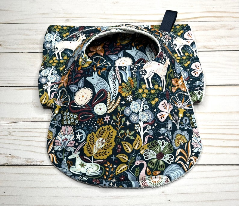 May include: A dark blue fabric bib with a white scalloped edge. The bib features a whimsical woodland pattern with deer, foxes, and birds in shades of white, brown, and green.