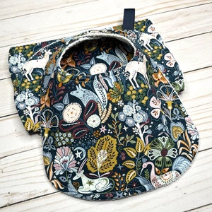 May include: A dark blue fabric bib with a white trim and a whimsical pattern of stylized animals, flowers, and stars. The pattern includes deer, foxes, birds, and floral motifs.