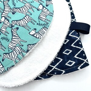 May include: A set of two baby burp cloths. One cloth is a light blue fabric with a white zebra print. The other cloth is a dark blue fabric with a white geometric pattern. The cloths are layered on top of each other.