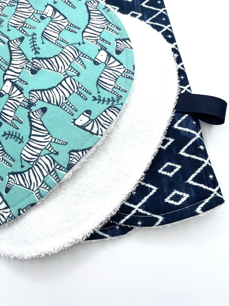 May include: A set of two baby burp cloths. One cloth is a light blue fabric with a white zebra print. The other cloth is a dark blue fabric with a white geometric pattern. The cloths are layered on top of each other.