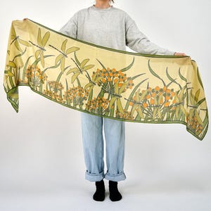 May include: A long, rectangular scarf with a light yellow background and a repeating pattern of green dragonflies and orange and green floral elements. The scarf has a green border.
