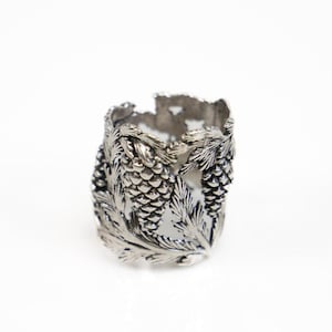 May include: A silver-colored ring featuring a detailed design of pine cones and leaves. The ring has an open, textured band with the pine cone and leaf motif wrapping around the circumference. The ring is set against a white background.